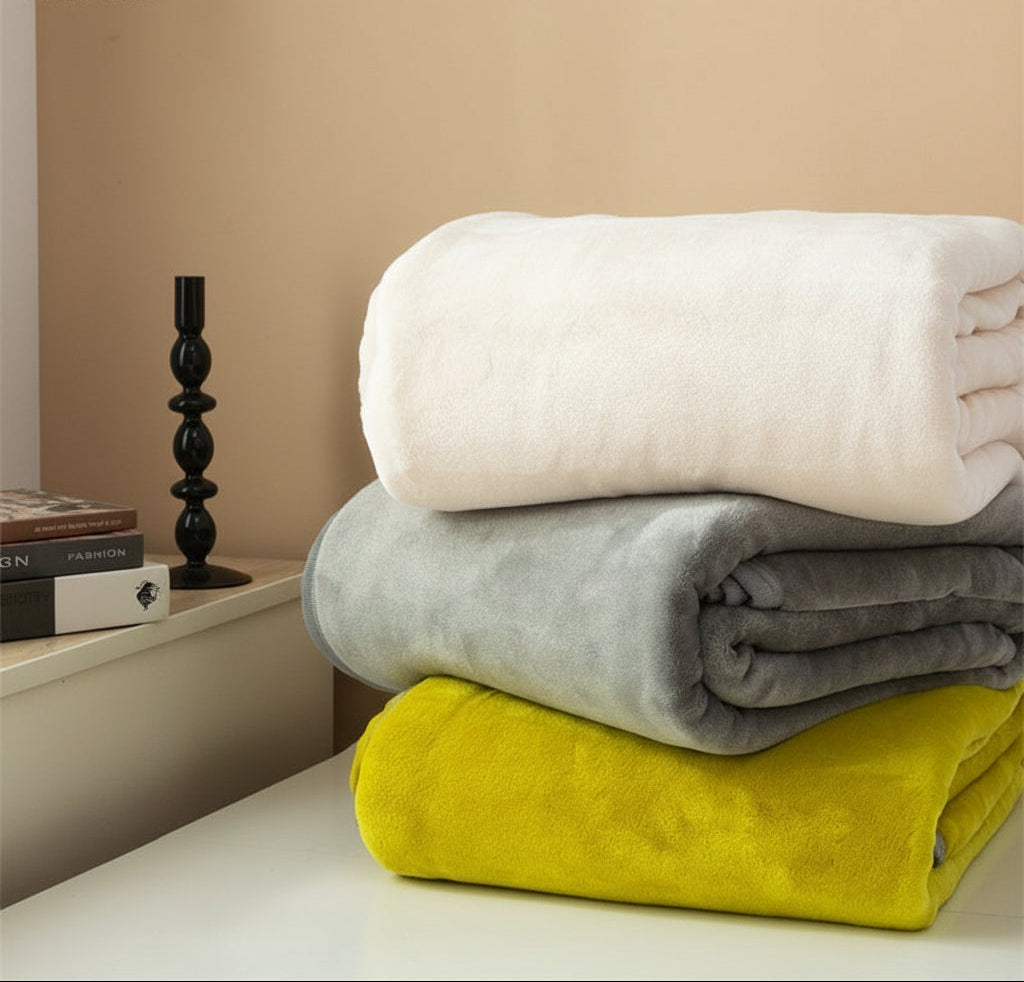 Stack of folded blankets in white, gray, and yellow on a surface with a candle and books in the background.