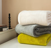 Stack of folded blankets in white, gray, and yellow on a surface with a candle and books in the background.